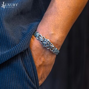 May include: A silver bracelet with a decorative design featuring a fleur-de-lis on a shield. The bracelet is worn on a person's wrist.