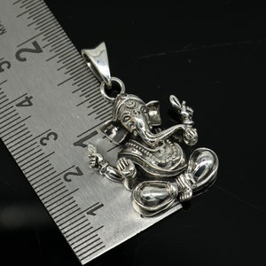 May include: Silver pendant featuring a detailed depiction of the Hindu deity Ganesha. The pendant is approximately 1.75 inches tall and is shown next to a ruler for scale.