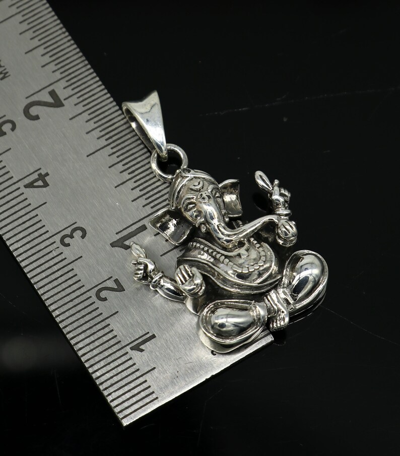 May include: Silver pendant featuring a detailed depiction of the Hindu deity Ganesha. The pendant is approximately 1.75 inches tall and is shown next to a ruler for scale.
