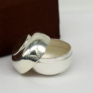 May include: A silver, round, lidded container with a smooth, shiny surface. The lid is slightly domed and the container is slightly wider at the top than the bottom.