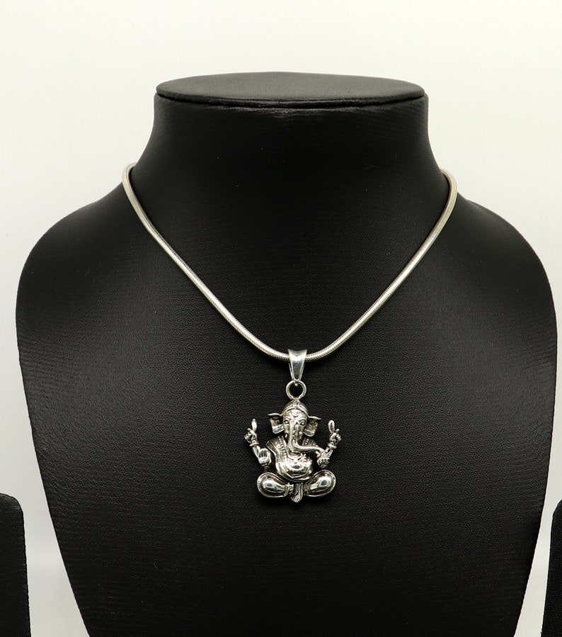 May include: A silver chain necklace with a pendant featuring a detailed depiction of the Hindu deity Ganesha. The pendant is a three-dimensional representation of Ganesha sitting in a meditative pose.