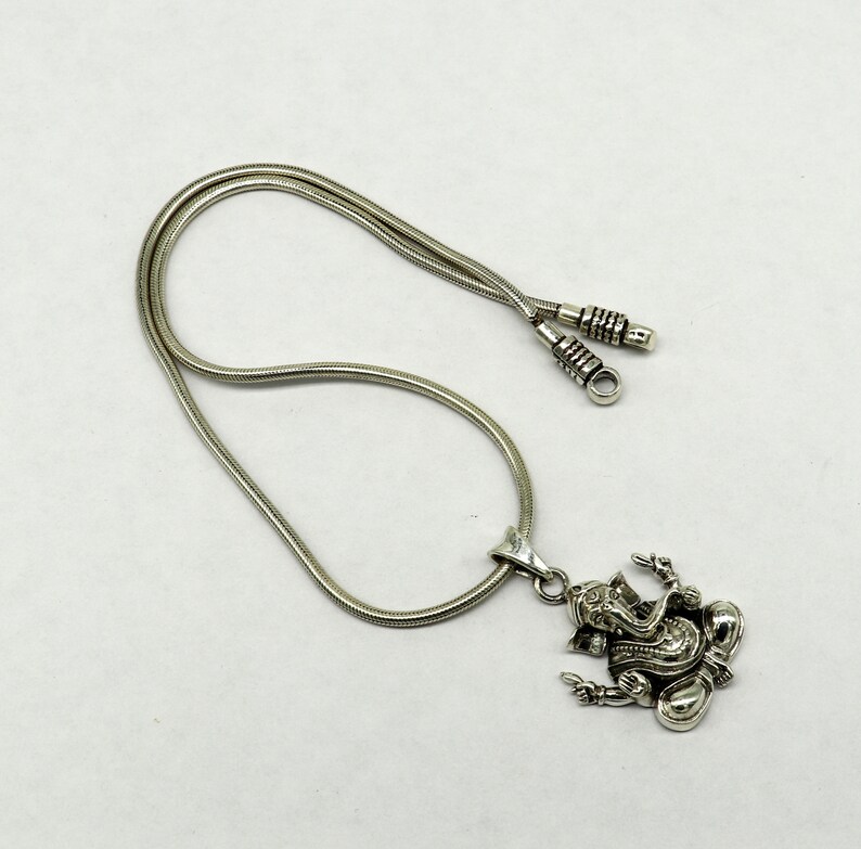 May include: A silver chain necklace with a silver pendant featuring a detailed depiction of the Hindu deity Ganesha. The chain is a snake chain style.