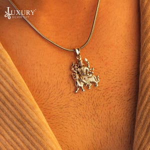 Può includere: Collana d'argento con un ciondolo che raffigura la dea indù Durga che cavalca un leone. Il ciondolo è dettagliato e intricato, con Durga che tiene armi nelle sue mani.