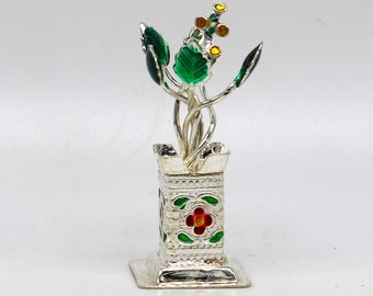 Op de afbeelding: Een zilveren vaas met een groen en geel bloemenontwerp. De vaas heeft een vierkante basis en een rechthoekig lichaam met een bloemenpatroon. De bloemen zijn van metaal en hebben een groene en gele emaille afwerking.