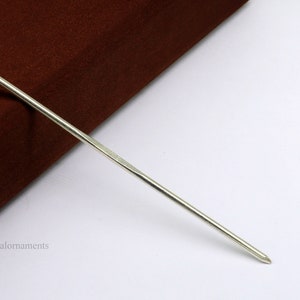 Puede incluir: Un palito para el pelo de plata, de aproximadamente 25 cm de largo, con un extremo ligeramente redondeado. El palito está sobre una superficie blanca.
