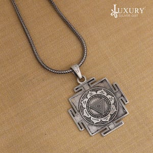 May include: A silver pendant necklace. The pendant has a square shape with a central triangle design and detailed circular patterns. The silver chain supports the pendant. The text "Luxury Silver Gift" is in the upper right corner.