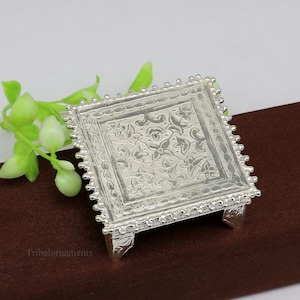 Puede incluir: Un pedestal cuadrado de plata con intrincados diseños florales y un borde de cuentas. El pedestal tiene cuatro patas y está hecho de metal.