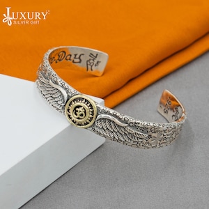 Puede incluir: Pulsera de brazalete de plata con un símbolo Om y alas. La pulsera está grabada con las palabras "Live Days Full" y otras frases inspiradoras.