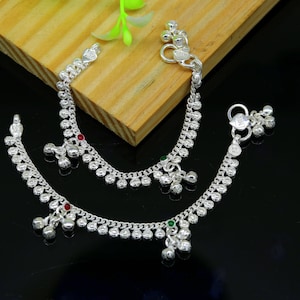 May include: Two silver anklets with small bells and green and red gemstones. The anklets are on a wooden surface.
