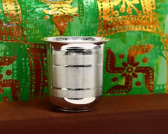 Puede incluir: Un vaso de beber de plata con un diseño grabado decorativo. El vaso está sobre una superficie marrón delante de una tela estampada verde y dorada.
