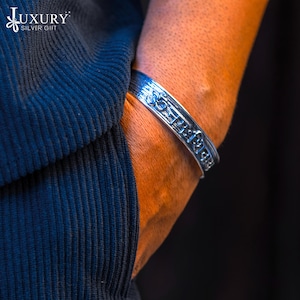 May include: A silver bracelet with an inscription and an Om symbol. The bracelet is worn on a person's wrist.