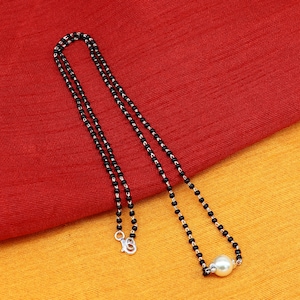 Pode incluir: Um colar de contas pretas com um único pingente de pérola branca. O colar está sobre um fundo de tecido vermelho e amarelo.