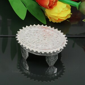 Puede incluir: Un soporte decorativo de plata con una parte superior redonda y tres patas. La parte superior del soporte tiene un diseño floral.