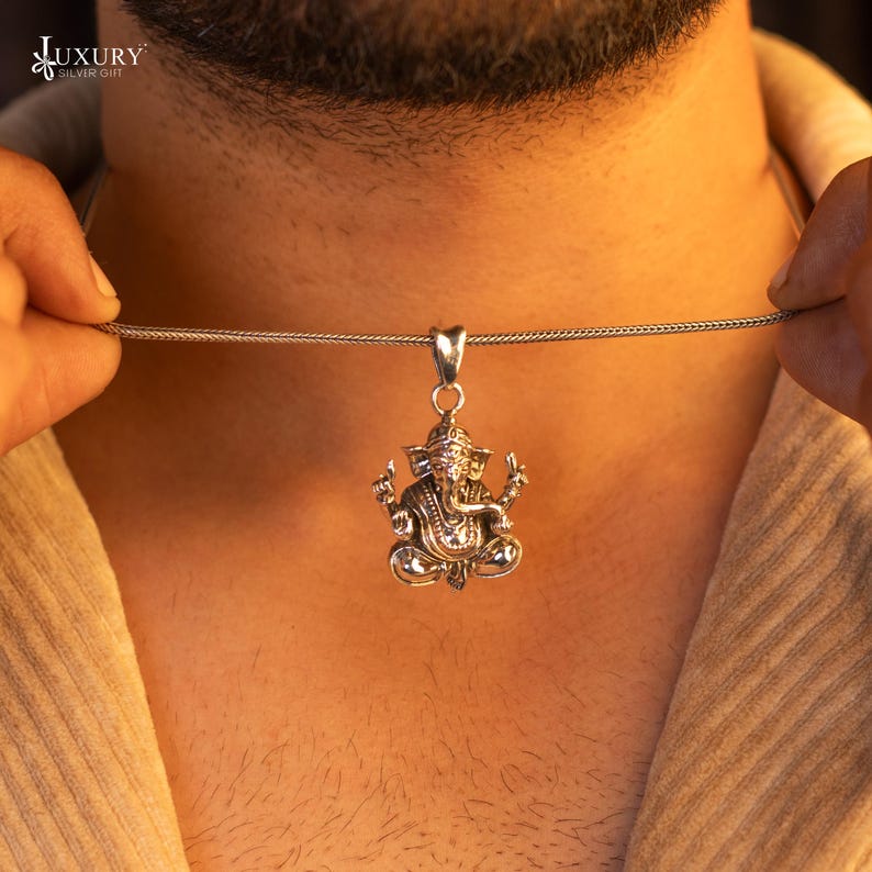 May include: A silver chain necklace with a detailed silver pendant of the Hindu deity Ganesha. The pendant is intricately designed with a sitting Ganesha with a trunk, large ears, and a crown.