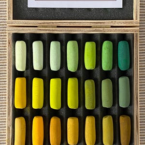 May include: A wooden box containing twenty-one rectangular pastel sticks arranged in three rows of seven, featuring a gradient of yellow, orange, and green shades. The lid interior displays a white card with the text: THIS BOX IS NUMBER _ OF A LIMITED EDITION OF _ BOXES. THIS PASTEL SET WAS HANDCRAFTED ON _ OF YEAR _.