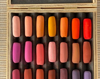 May include: A wooden box containing twenty-one rectangular pastel sticks arranged in three rows. The pastels feature a gradient of warm colours including pink, orange, red, brown, and purple. A white label inside the lid includes the text: THIS BOX IS NUMBER ___ OF A LIMITED EDITION OF ___ BOXES. THIS PASTEL SET WAS HANDCRAFTED ON ___ OF YEAR ___.