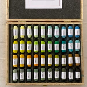 May include: A wooden box filled with 48 pastel sticks in various shades of green, yellow, and blue. The sticks are labeled "MAMMUT".