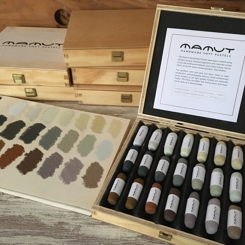 以下が含まれることがあります： 木製の箱に、手作りのソフトパステルが24本入っています。パステルは、行に並べて配置され、「Mamut Handmade Soft Pastels」とラベル付けされています。箱は明るい木でできており、黒いラベルには「Mamut Handmade Soft Pastels」というテキストと、パステルの説明が書かれています。