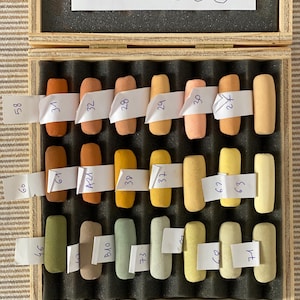 May include: A wooden box containing twenty-one rectangular pastel sticks arranged in three rows, each labeled with a white paper strip featuring handwritten numbers. The colors range from earthy browns and oranges to yellows, greens, and muted grays.