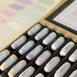 Może przedstawiać: Drewniane pudełko wypełnione pastelowymi pastelami. Każdy patyczek pasteli ma białą etykietę z nazwą marki "MAMUT Handmade Soft Pastels". W tle widoczna jest paleta kolorów z próbkami.