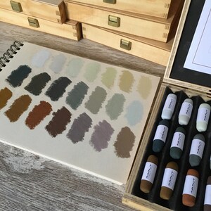 以下が含まれることがあります： 木製の箱に入った24本のパスツールスティックのセット。パスツールは、茶色、灰色、緑色の色見本を並べた列に配置されています。箱は木でできており、黒い内装になっています。