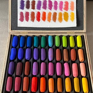 Peut inclure: Une boîte en bois contenant un ensemble de bâtons de pastel rectangulaires disposés en rangées par couleur, incluant des nuances de bleu, vert, violet, rouge, orange et jaune. Une carte d'échantillons en papier blanc est fixée au couvercle.