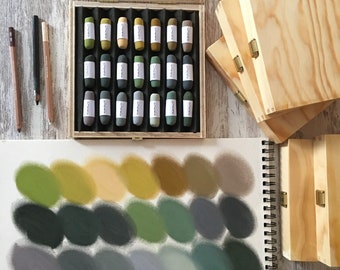 May include: A set of 24 oil pastels in a wooden box. The pastels are arranged in rows of four, with each row containing a different shade of green. The pastels are labelled with their colour names.