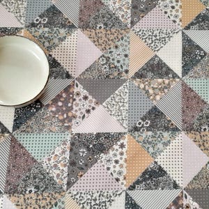 Op de afbeelding: Een decoratieve tafelloper met een geometrisch patchwork-ontwerp in tinten grijs, beige en lichtblauw. Een witte keramische kom met een zwarte rand ligt op de loper. Het patroon omvat bloemen-, gestreepte en gestippelde stoffen.