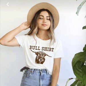 Może przedstawiać: Biała koszulka z brązową ilustracją krowy szkockiej górskiej i nadrukiem "BULL SHIRT" z przodu.