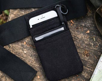 May include: A black fabric belt pouch featuring a wide elastic strap, a vertical pocket containing a white smartphone, and a smaller front pocket holding a stack of cards. A black carabiner is attached to the top edge of the pouch.