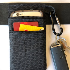 May include: Black fabric phone holder with a small pocket for a credit card. The holder is attached to a black elastic band with a carabiner clip and keys.
