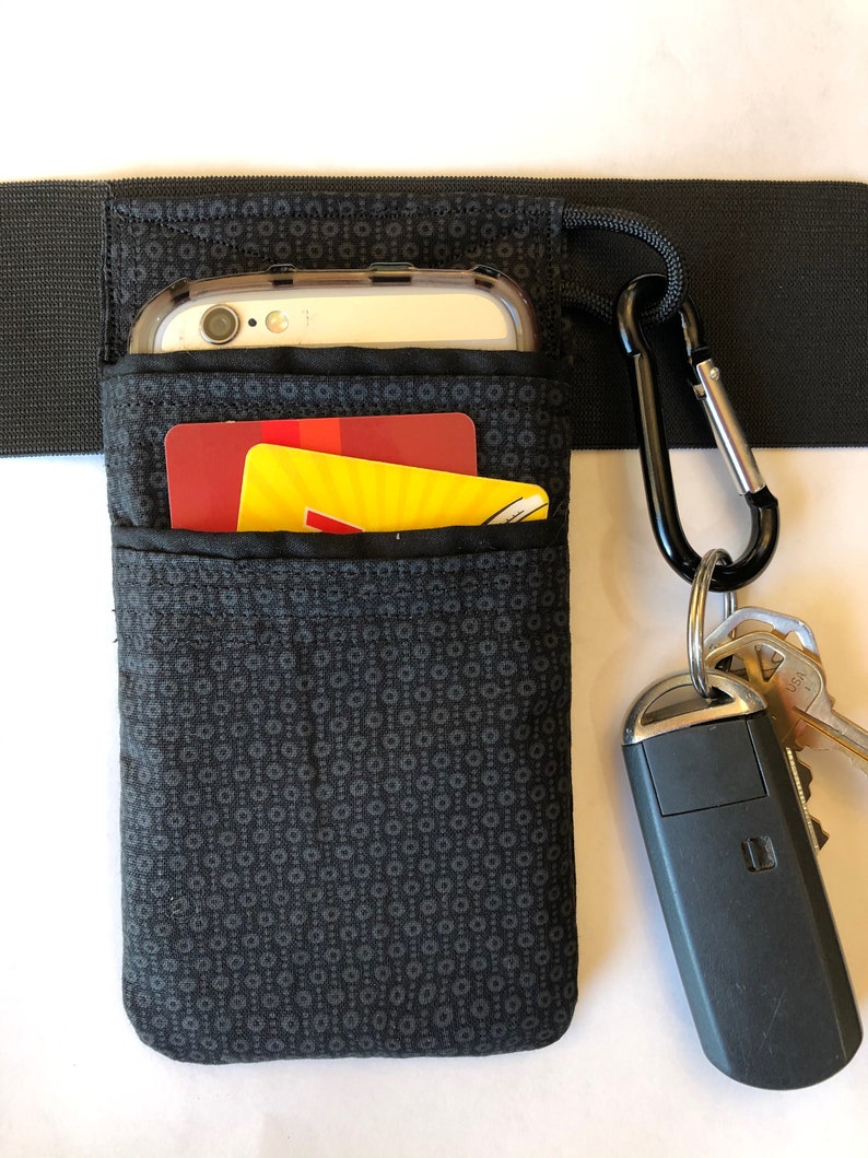 May include: Black fabric phone holder with a small pocket for a credit card. The holder is attached to a black elastic band with a carabiner clip and keys.