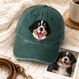 May include: A dark green baseball cap featuring an embroidered Bernese Mountain Dog face on the front panel. The dog has black, brown, and white fur with a pink tongue visible. The cap has a curved brim and a structured crown with stitched eyelets.