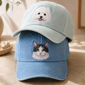 May include: A light blue denim baseball cap with an embroidered white Bichon Frise dog face sits atop a medium blue denim baseball cap featuring an embroidered grey and white cat face. Both caps have a curved brim and a structured crown design.