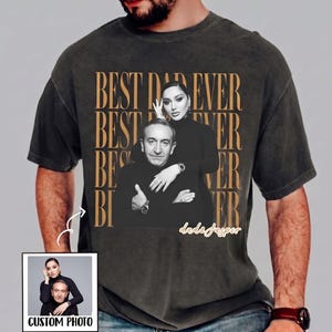 以下が含まれることがあります： 男性と女性の白黒写真がプリントされた濃いグレーのTシャツ。「BEST DAD EVER」の文字が金色のパターンで繰り返されています。「CUSTOM PHOTO」という文字が入った小さな画像も表示されています。
