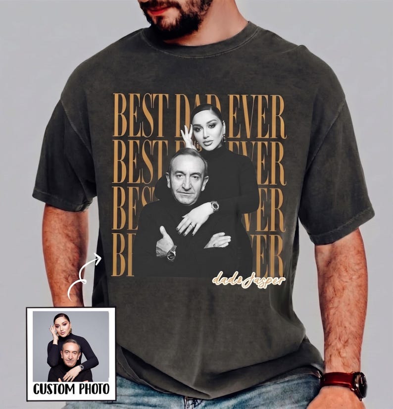 以下が含まれることがあります： 男性と女性の白黒写真がプリントされた濃いグレーのTシャツ。「BEST DAD EVER」の文字が金色のパターンで繰り返されています。「CUSTOM PHOTO」という文字が入った小さな画像も表示されています。