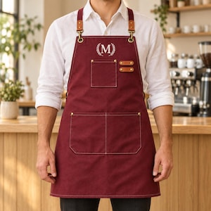 Könnte beinhalten: Eine tiefburgunderrote Schürze mit einem weißen, gestickten Buchstaben M in einem Lorbeerkranz. Die Schürze verfügt über eine Brusttasche, zwei große untere Taschen und braune Lederriemen mit messingfarbenen Beschlägen und Nieten.