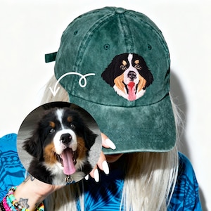 May include: A faded green baseball cap with an embroidered Bernese Mountain Dog design. The dog's face is black, brown, and white with a pink tongue. The cap has a curved peak and an adjustable strap at the back.