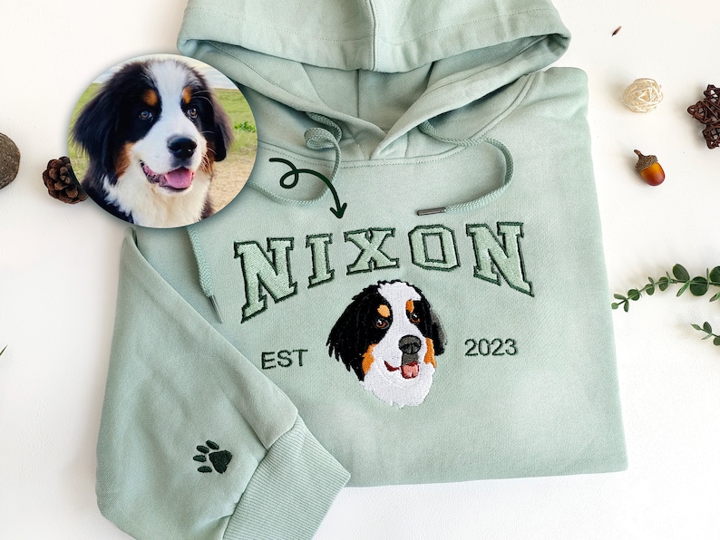 K&ouml;nnte beinhalten: Ein hellgr&uuml;ner Kapuzenpullover mit einem Pfotenabdruck auf dem &Auml;rmel. Der Pullover hat ein gesticktes Design mit dem Namen "Nixon" in einer College-Schriftart, dem Jahr "2023" und einem gestickten Bild eines Berner Sennenhundes.