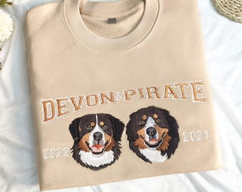 Könnte beinhalten: Beiges Sweatshirt mit goldfarbener Stickerei "DEVON & PIRATE". Darunter zwei Hundeporträts mit den gestickten Jahreszahlen 2022 und 2021. Die Hunde sind schwarz, braun und weiß.