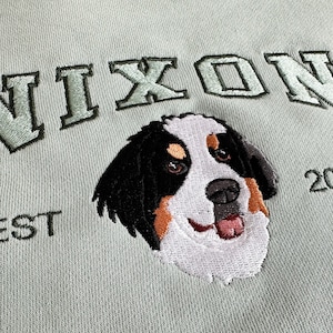 K&ouml;nnte beinhalten: Gr&uuml;nes Sweatshirt mit gesticktem Text "NIXON EST 2023" und einem Berner Sennenhund.