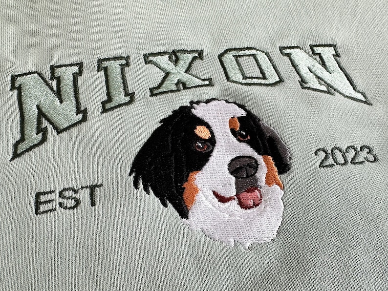 K&ouml;nnte beinhalten: Gr&uuml;nes Sweatshirt mit gesticktem Text "NIXON EST 2023" und einem Berner Sennenhund.