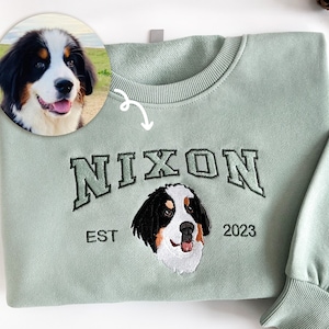 K&ouml;nnte beinhalten: Ein hellgr&uuml;ner Pullover mit einem gestickten Hundeportr&auml;t in Schwarzwei&szlig;, dem Text "NIXON EST 2023" und einem Pfotenabdruck.