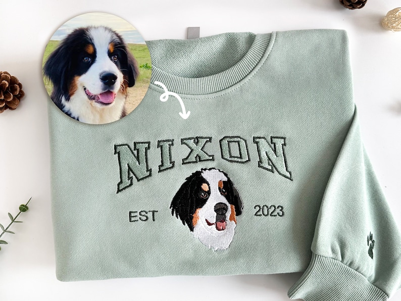 K&ouml;nnte beinhalten: Ein hellgr&uuml;ner Pullover mit einem gestickten Hundeportr&auml;t in Schwarzwei&szlig;, dem Text "NIXON EST 2023" und einem Pfotenabdruck.
