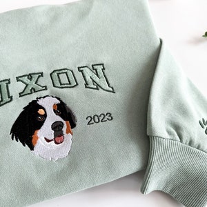 K&ouml;nnte beinhalten: Ein hellgr&uuml;ner Pullover mit dem Wort "NIXON" in dunkelgr&uuml;n gestickt, darunter ein Berner Sennenhund, und darunter der Text "EST 2023". Auf dem rechten &Auml;rmel ist ein gr&uuml;ner Pfotenabdruck gestickt.