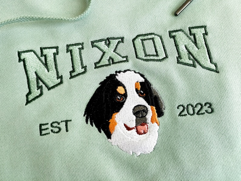 K&ouml;nnte beinhalten: Ein hellgr&uuml;ner Hoodie mit dem Wort "NIXON" in Gr&uuml;n gestickt, einem Hundekopf in Wei&szlig;, Schwarz und Braun gestickt und dem Text "EST 2023" in Gr&uuml;n gestickt.