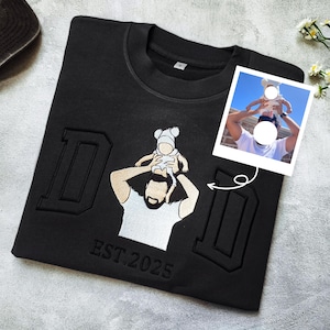 Peut inclure: Sweat-shirt noir avec broderie "DAD" et un dessin d'une personne portant un bébé. Le sweat-shirt comprend le texte "EST. 2025" et une petite photo. Col rond.