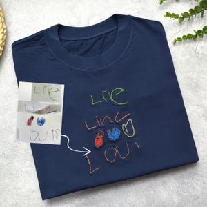 May include: A navy blue t-shirt with embroidered artwork. The design includes colourful text and drawings, including the name "Louis". A small drawing is shown next to the shirt, with a white arrow pointing to the shirt.