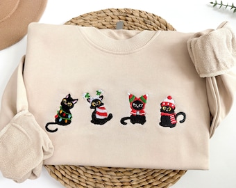 May include: A beige sweatshirt featuring embroidered black cats wearing festive holiday attire, including a Santa hat, reindeer antlers, and a Christmas tree ornament.