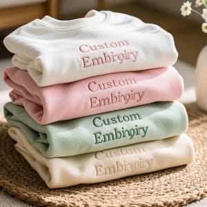 May include: A stack of four folded sweatshirts in cream, pink, mint green, and beige. Each sweatshirt has the words "Custom Embroidery" embroidered in a matching color. The sweatshirts are stacked on a woven mat.